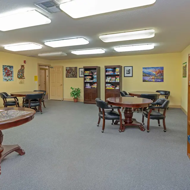 A spacious community room featuring several round tables with chairs, shelves filled with books and games, and artwork on the walls. The decor includes a light yellow wall color and bright overhead lighting.