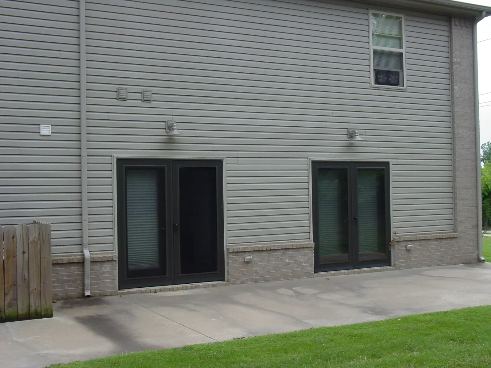 Two black doors with glass panels are set into a gray, horizontal siding wall. A fenced area is visible on the left.