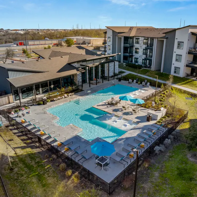 Aerial view of a modern apartment complex featuring a large swimming pool, lounge chairs, and a deck area surrounded by greenery.