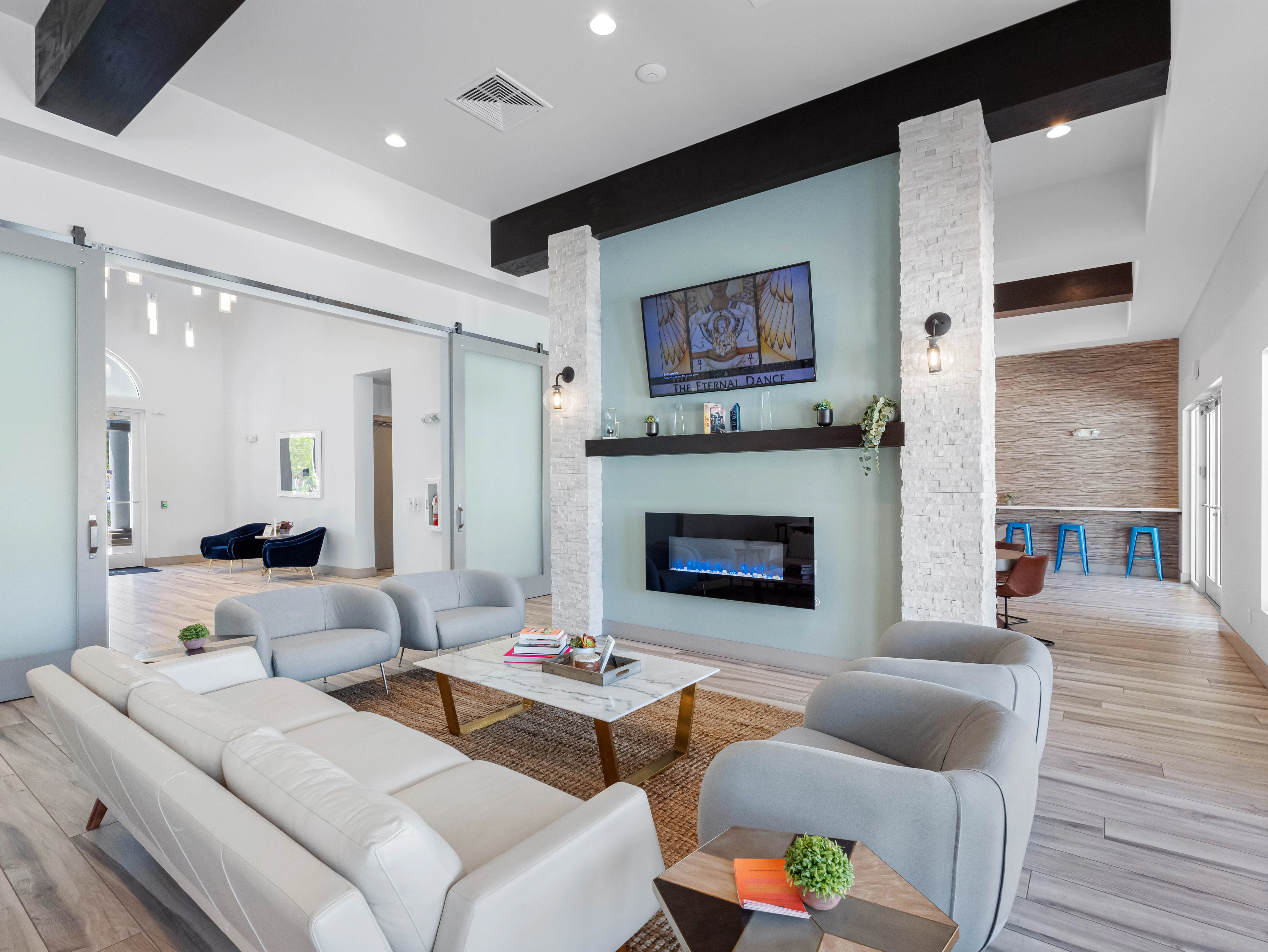 A modern living room featuring a white leather sectional sofa, a sleek coffee table, and a stone fireplace. There is a television mounted on the wall above the fireplace, and the room has large windows allowing natural light. Adjacent to the living area is a glimpse of another space with blue stools and wooden elements.