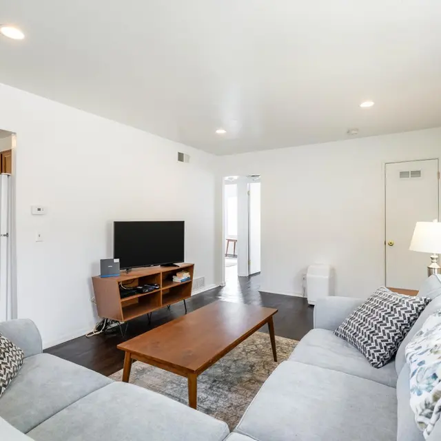 A bright and airy living room featuring a gray sectional sofa, a wooden coffee table, and a television on a stand. The walls are white, and natural light streams in through windows.