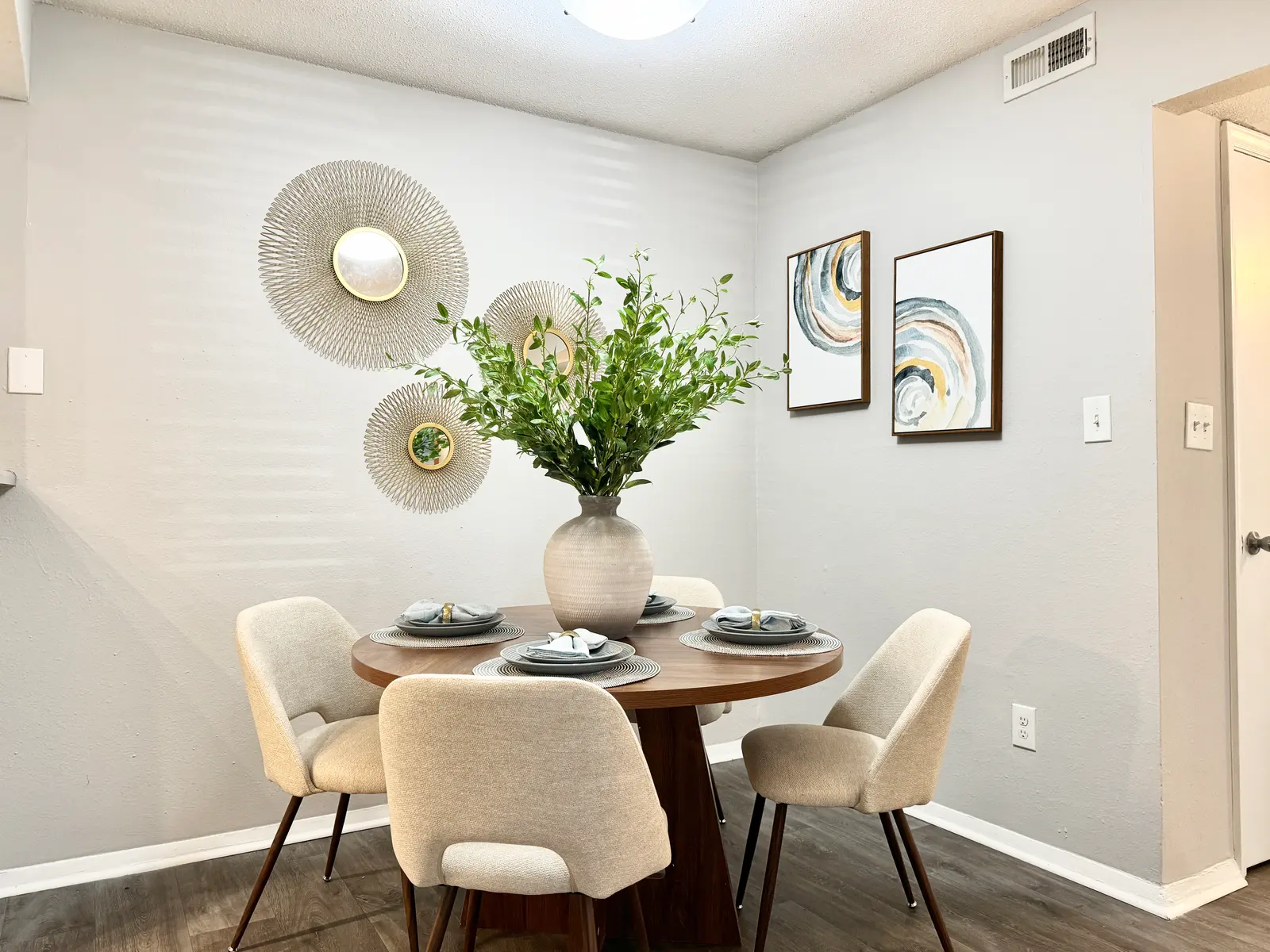 A modern dining area featuring a round wooden table with four cream-colored upholstered chairs. A large vase with greenery is the centerpiece. The walls are adorned with decorative plates and abstract art in frames. The room has soft lighting and a warm, inviting atmosphere.
