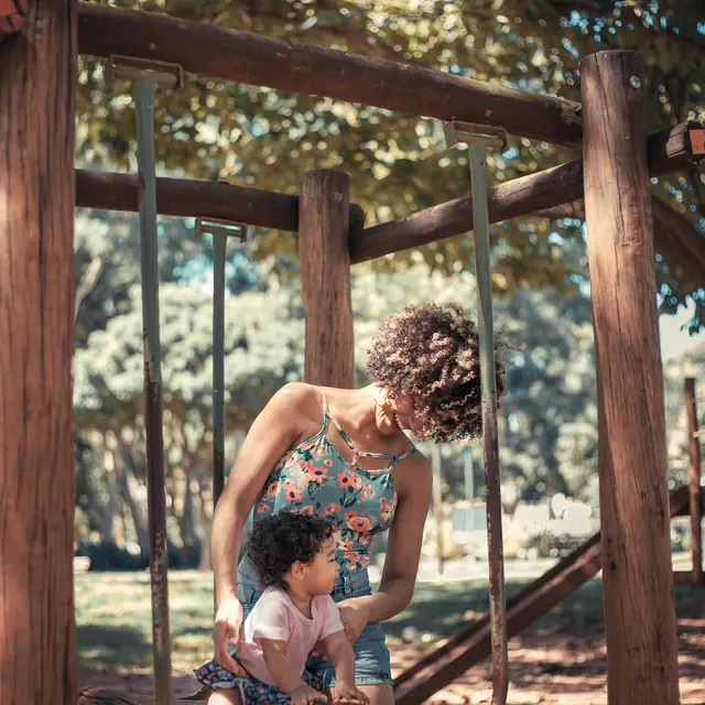 Mother and Daughter at the Playground A woman in a floral dress is helping a small child on a swing in a park. The sun filters through the trees, creating a warm atmosphere.
