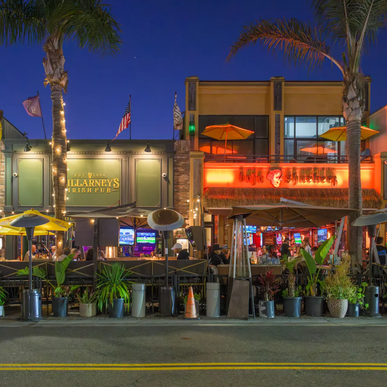 A vibrant street scene featuring a restaurant with outdoor seating on a busy night. The area is lined with palm trees and decorated with umbrellas and lights. Several people can be seen enjoying their meals outside, with the restaurant’s sign glowing prominently.