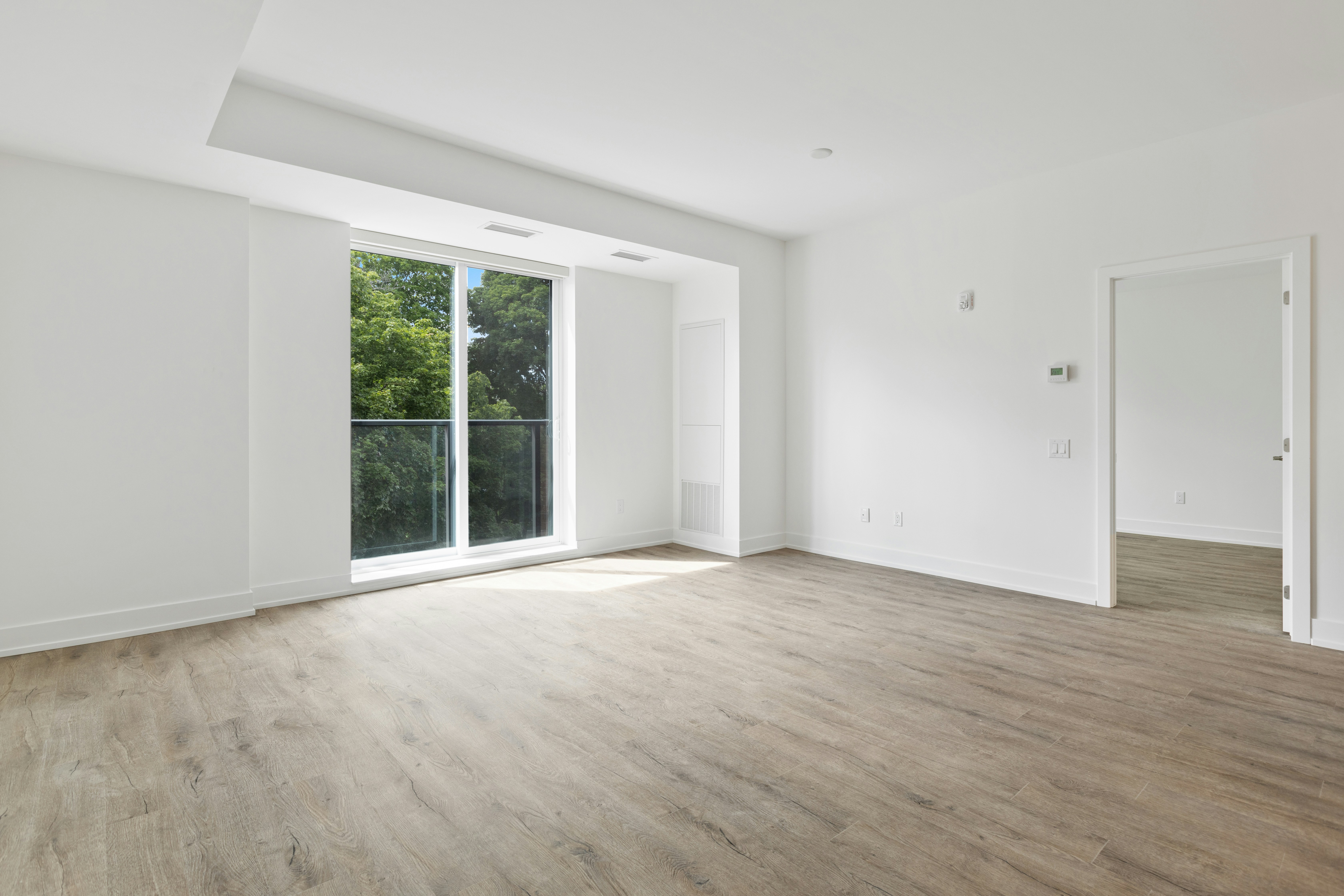A spacious, empty room with large windows and light-colored walls. The room features modern flooring and a door leading to another space.