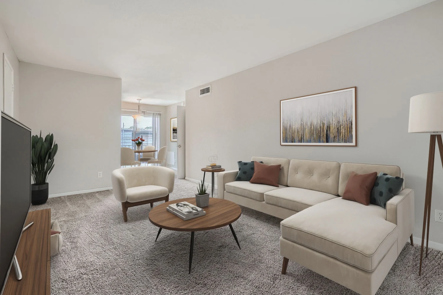 A modern living room featuring a light beige sectional sofa with colorful pillows, a round wooden coffee table, and a stylish lamp. The room has light-colored walls and a cozy carpet, with a decorative piece of art on the wall.