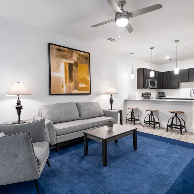 A modern living room with a gray sofa and armchairs on a blue rug, featuring a coffee table. The area has decorative lamps and an abstract painting on the wall. The kitchen area is in the background with dark cabinets and a white countertop.