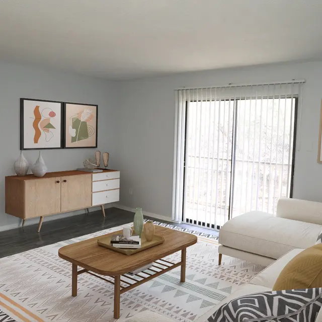 A modern living room featuring a light color palette, a white sectional sofa, a wooden coffee table, and a decorative area rug. There are two pieces of abstract artwork on the wall, a sideboard with decorative items, and large sliding glass doors leading to a balcony.