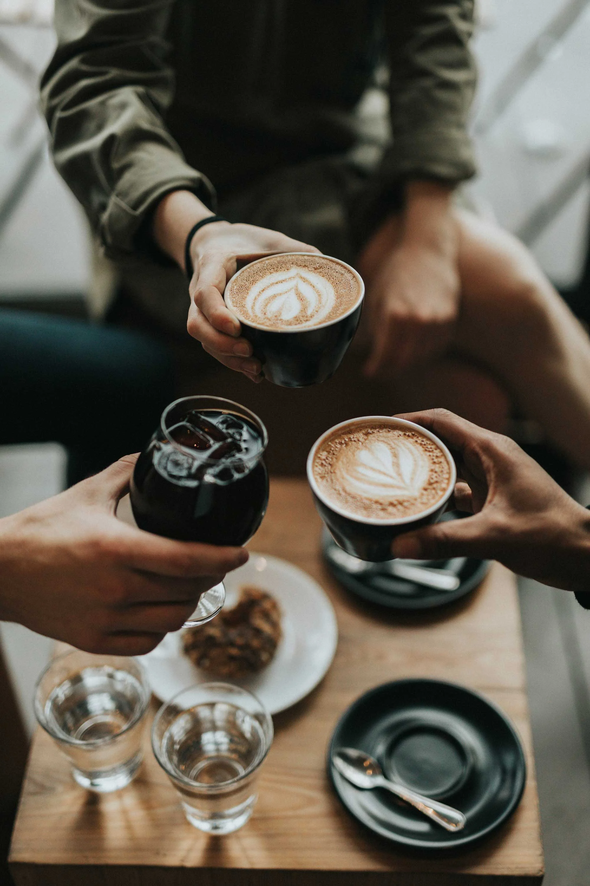 A close-up view of hands holding coffee cups, with a wooden table displaying drinks and a dessert.
