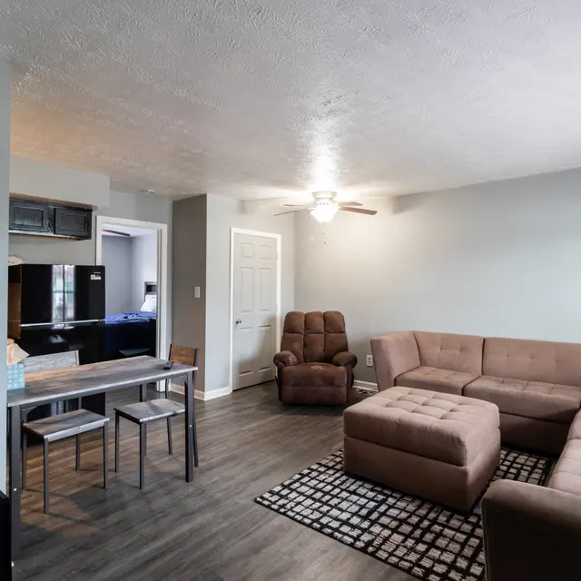 A cozy living room featuring a sectional sofa, a wooden dining table with chairs, and a recliner. The room has gray walls and a ceiling fan, with a patterned rug on the floor.