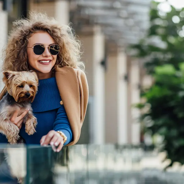 A smiling woman with curly hair, wearing sunglasses and a stylish outfit, holds a small dog while leaning against a glass railing. In the background, there are trees and modern architecture.