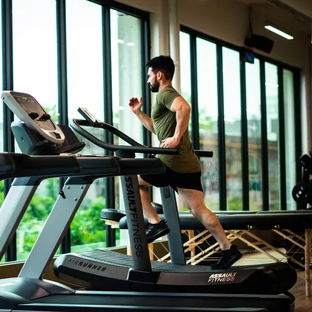 A man runs on a treadmill in a bright gym, surrounded by large windows and fitness equipment.