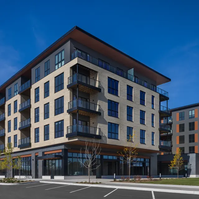 A modern multi-story apartment building featuring a mix of brick and glass architecture, with multiple balconies and a clear blue sky in the background.