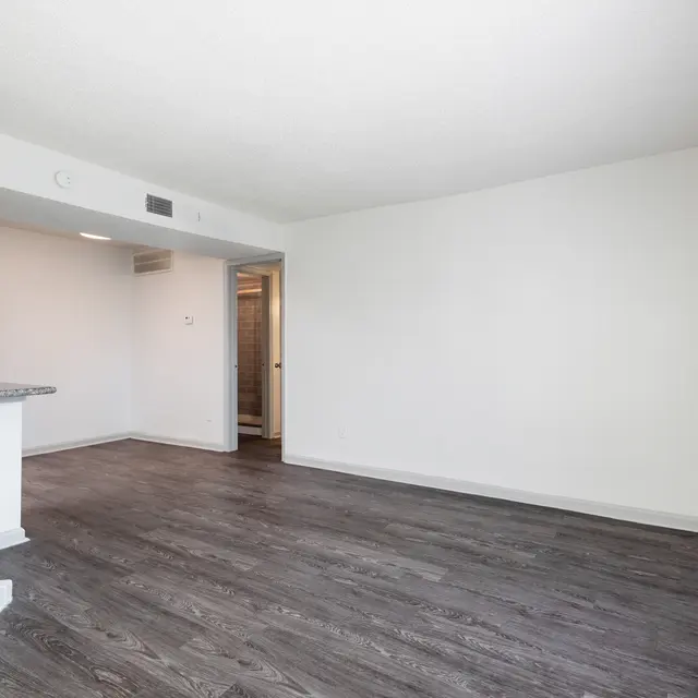 Empty living room with hardwood floors and white walls, featuring a small kitchen area and a doorway leading to another room.