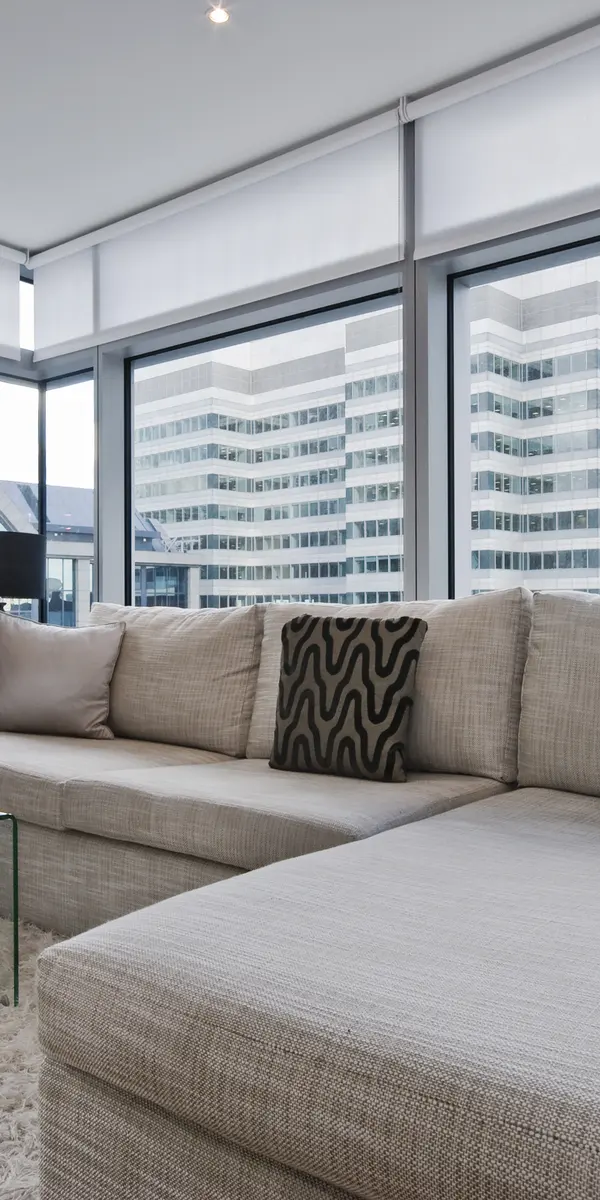 Modern living room with a cozy sectional sofa, a glass coffee table, and a television, featuring large windows with city views.