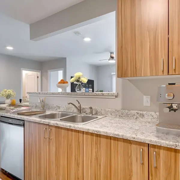 A contemporary kitchen with wood cabinetry, granite countertops, a double sink, and a stainless steel dishwasher. The counter has a coffee maker and is decorated with flowers and a bowl of fruit, creating a welcoming, stylish space. A dining area is visible in the background.
