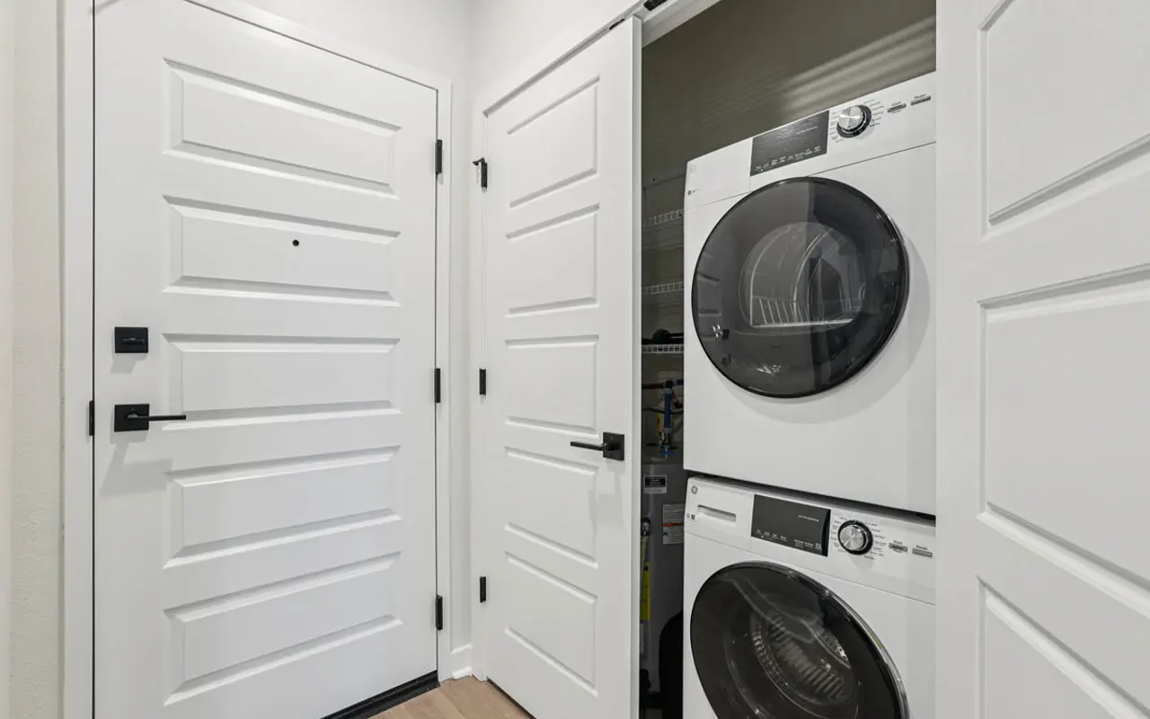 Compact Laundry Room near Entryway A modern laundry area featuring stacked washer and dryer units situated in a hallway near a front door. The walls are painted in a light color, and the flooring is a warm laminate. Two white doors lead into the laundry space, creating a compact design.