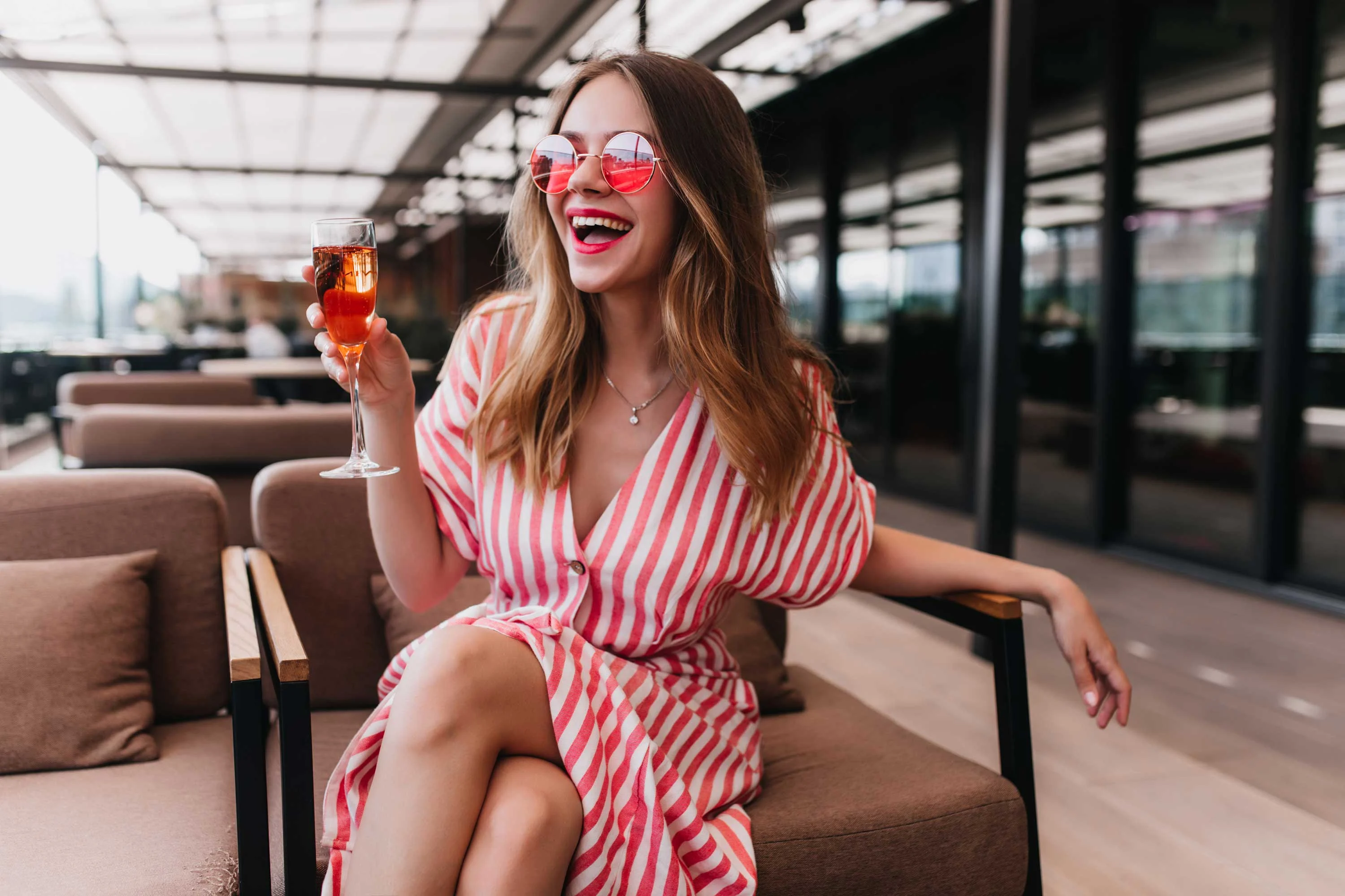 A woman in a striped dress sitting in a modern lounge area, holding a drink and smiling. She is wearing sunglasses and has long hair.