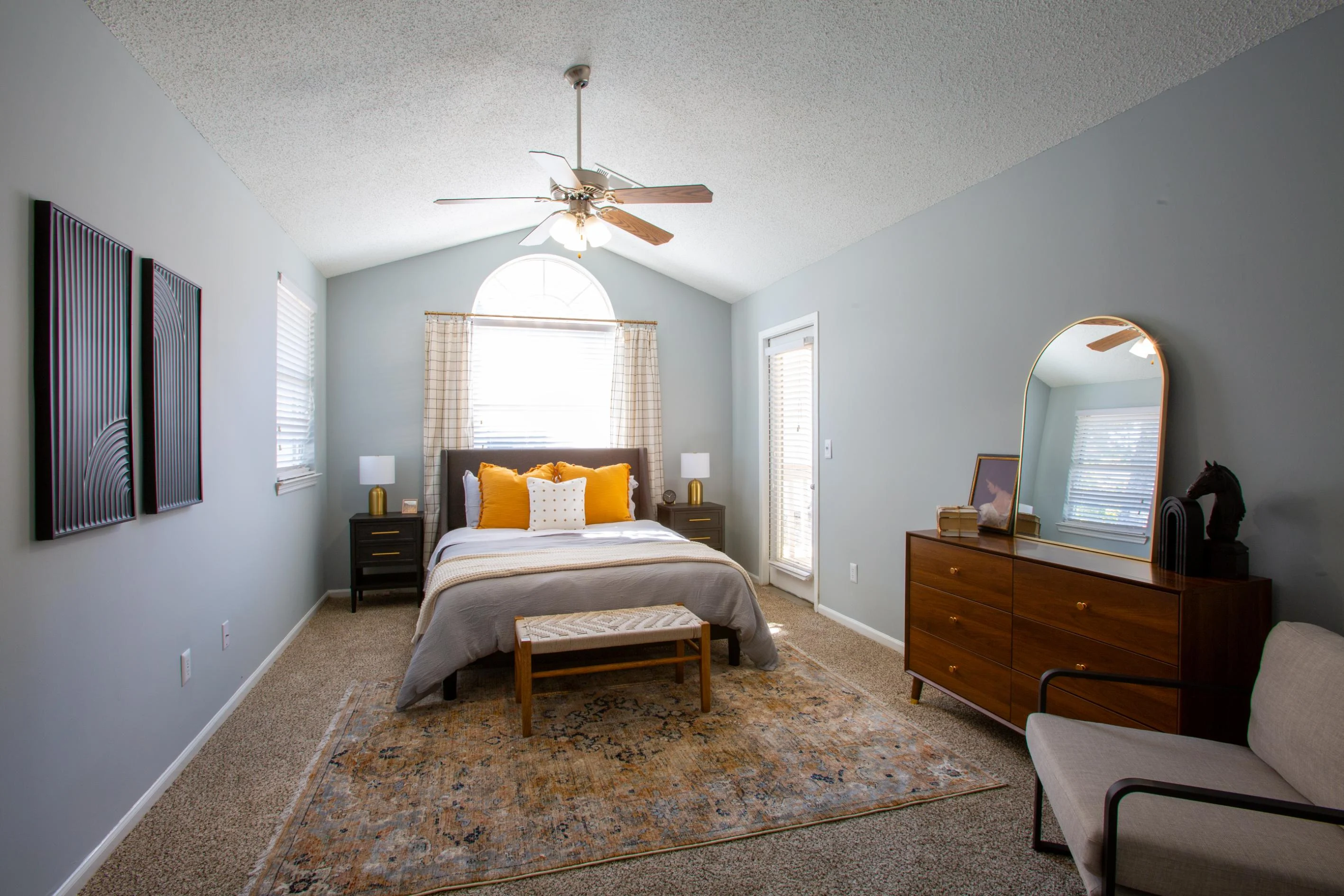 A beautifully decorated bedroom featuring a bed with white linens and decorative pillows, a wooden bench at the foot of the bed, and a dresser. Natural light floods the space through a large window with sheer curtains. The room has a light gray painted wall, a plush rug on the carpet, and modern decor elements like framed artwork and a round mirror.