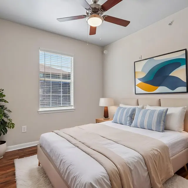 Bedroom (Staged) - Tigoni Villas A cozy bedroom featuring a queen-sized bed with decorative pillows, wooden nightstands, and a large window beside a potted plant. A wall-mounted TV and a ceiling fan are also visible.