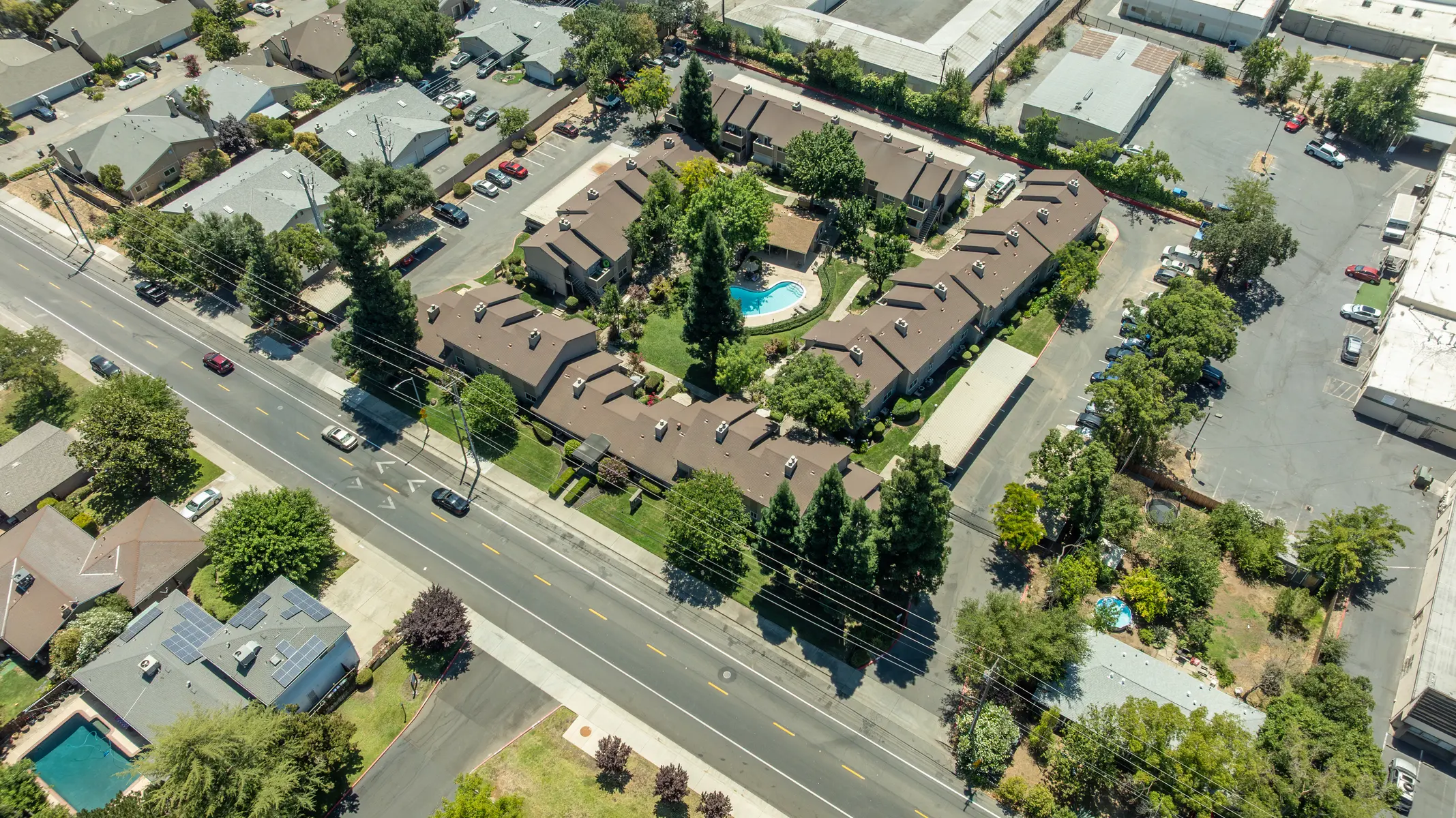 Aerial view of an apartment complex surrounded by trees, with a swimming pool visible, and single-family homes nearby along a road.