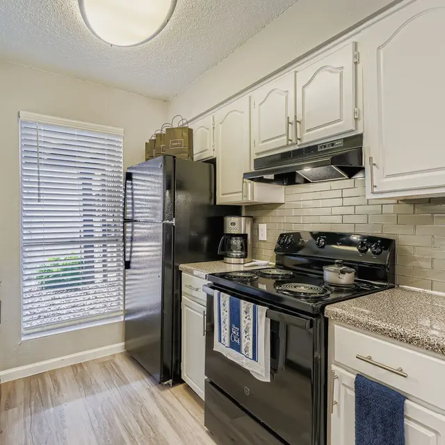 Stylish Kitchen Interior A modern kitchen featuring white cabinets, a black refrigerator, and a black stove. The area is well-lit with natural light coming through a window adorned with blinds. There is a granite countertop and stylish tile backsplash.