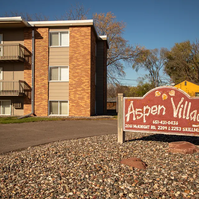 Exterior view of a multi-unit residential building with a sign reading 'Aspen Village' at the entrance, surrounded by gravel and landscaping.