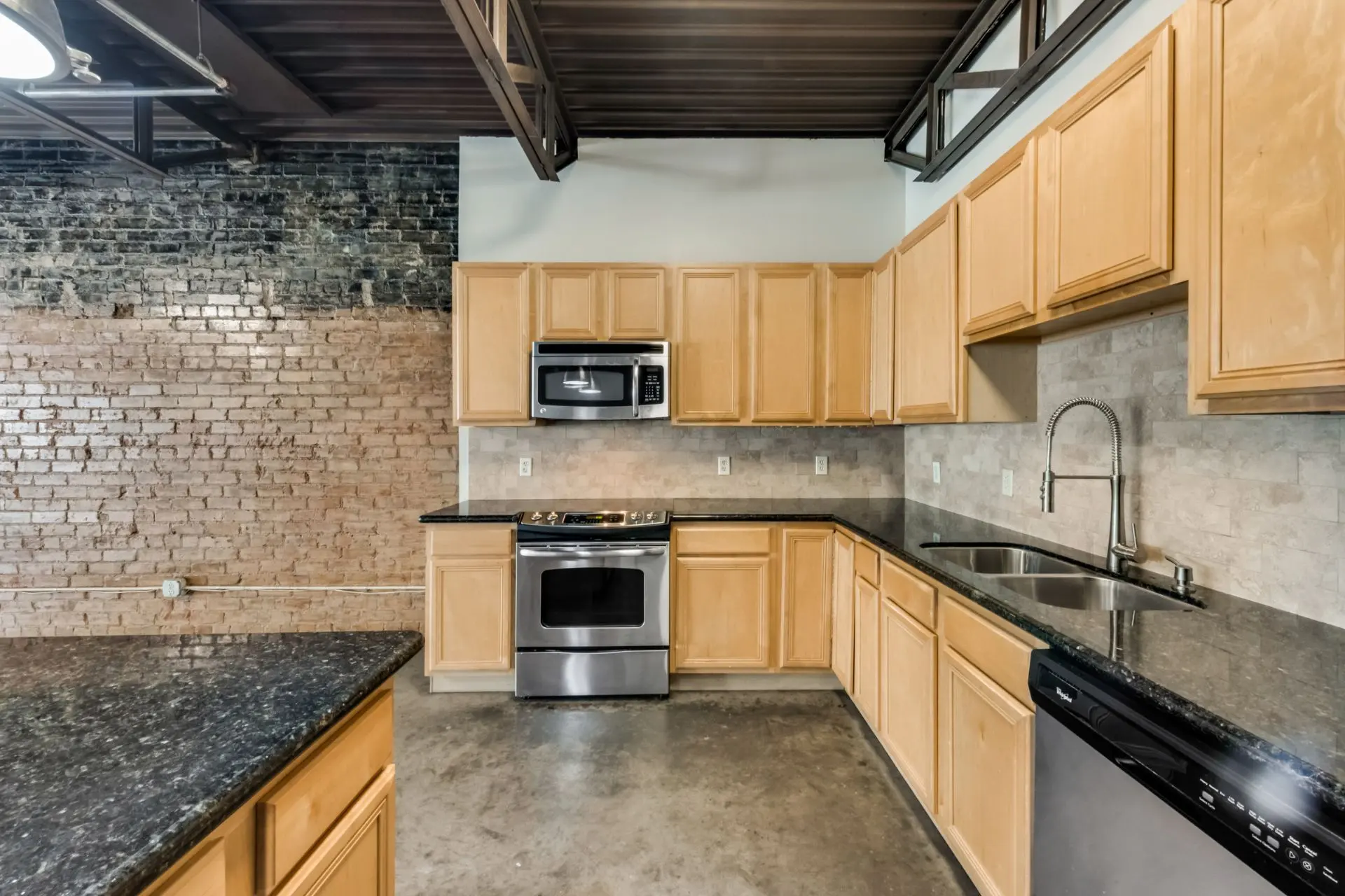 A renovated kitchen featuring light wooden cabinets, black countertops, stainless steel appliances, and a brick accent wall.
