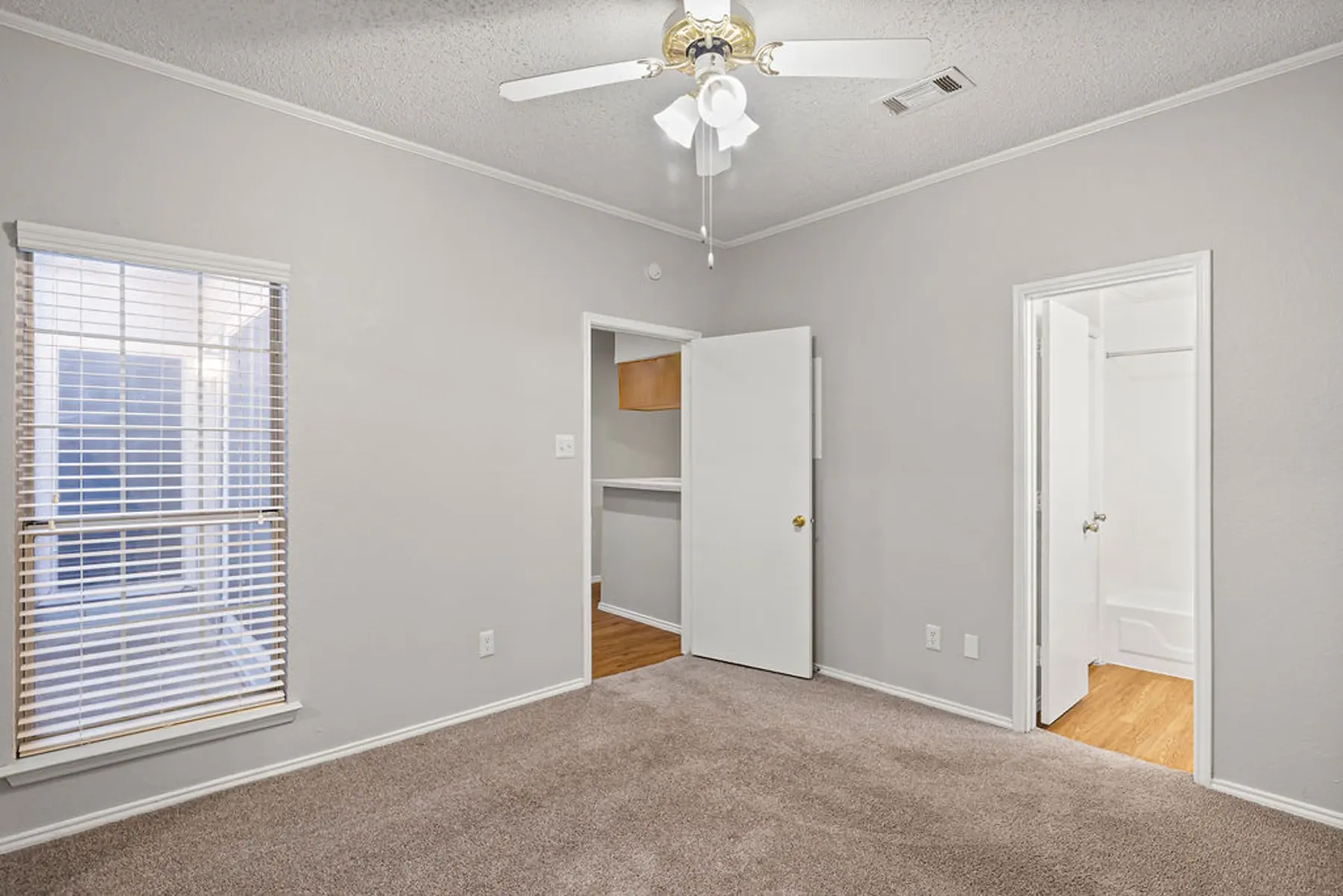 Empty Bedroom Interior A view of an empty bedroom with light gray walls and carpet flooring. The room features a ceiling fan, a window with blinds on the left, and two doorways leading to other spaces.