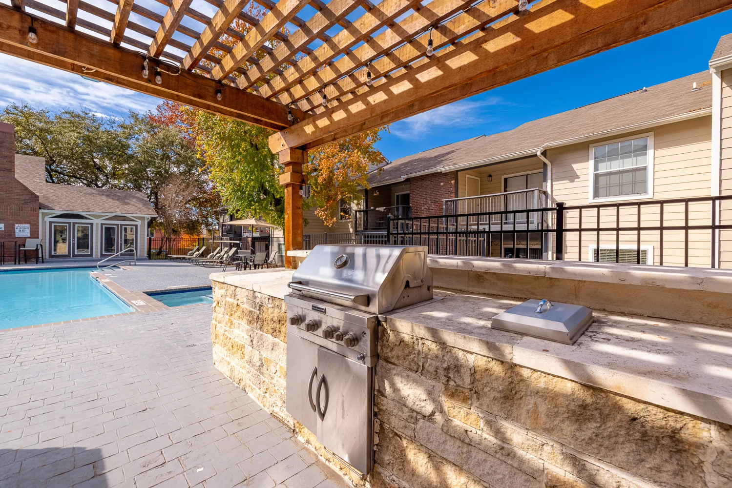 Barbecue area next to a swimming pool, featuring a grill under a wooden pergola with a clear blue sky in the background.