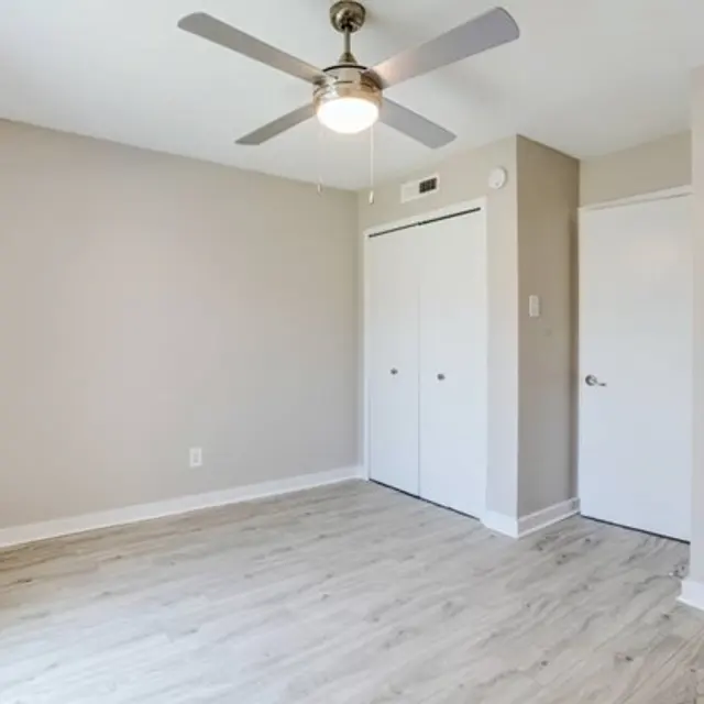 A minimalistic room featuring light wooden flooring and neutral-colored walls. There is a ceiling fan in the center and two white closet doors on one wall, with an additional closed door on the right. The room is well-lit with natural light coming from the window.