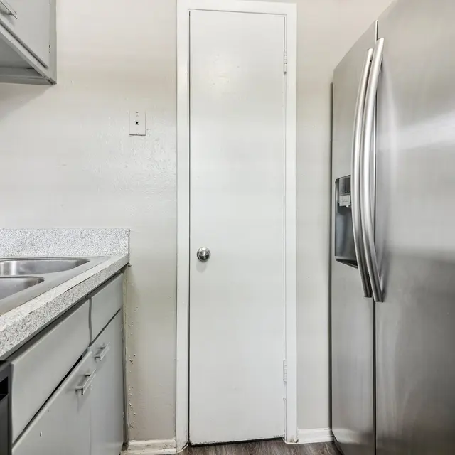 A modern kitchen with stainless steel appliances and a door leading to another room. The kitchen features a double sink, gray countertop, and cabinets above. The walls are painted in a light color, and the flooring appears to be laminate.