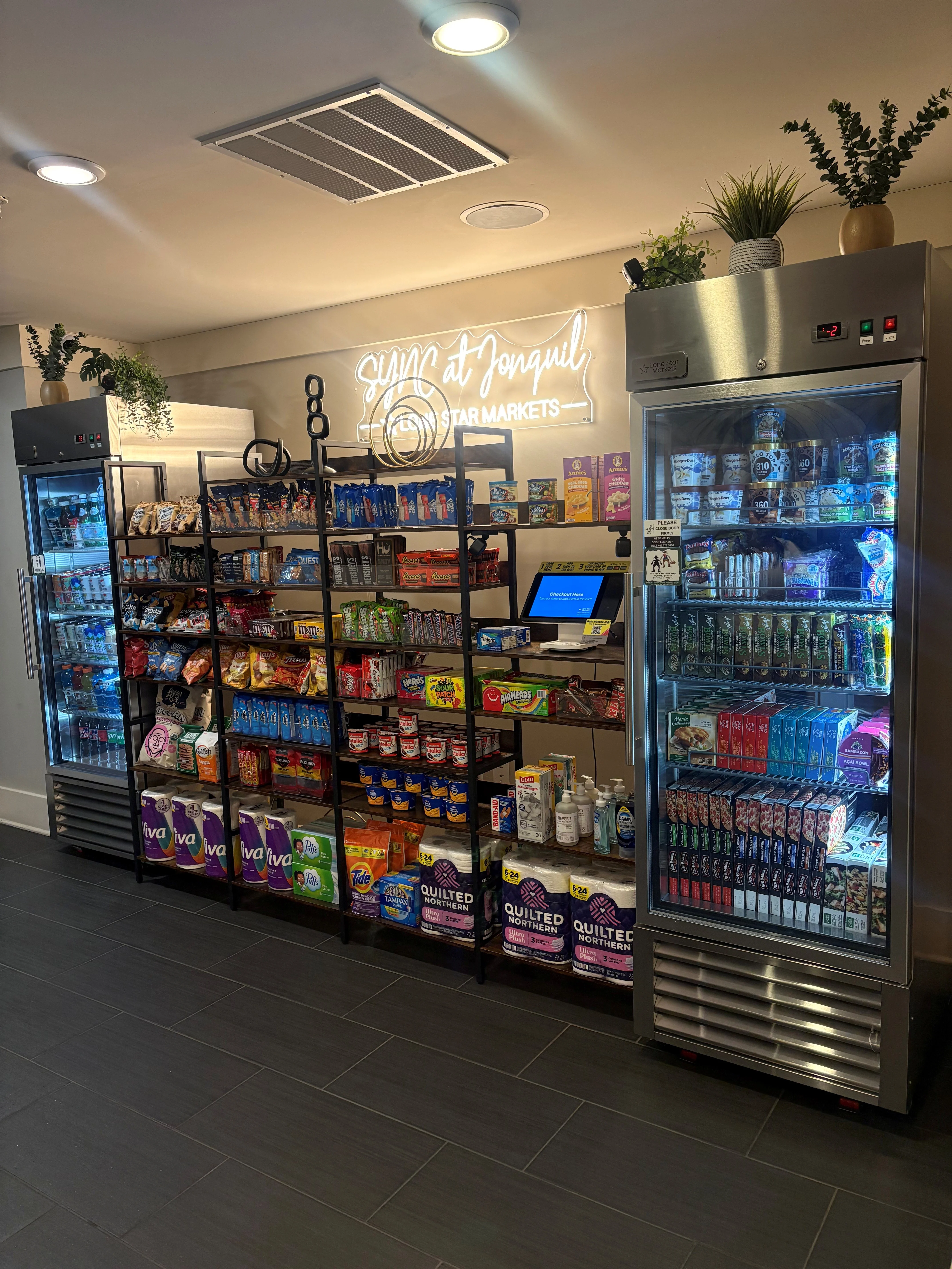 Self-Service Market Area A well-organized self-service market area with glass-front coolers and shelves filled with snacks and drinks, featuring a digital checkout station.