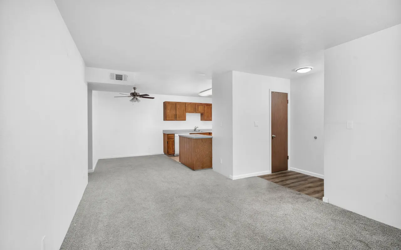 Spacious living area in an apartment with light-colored walls and plush carpet, featuring a ceiling fan. The area connects to a kitchen with wooden cabinets.