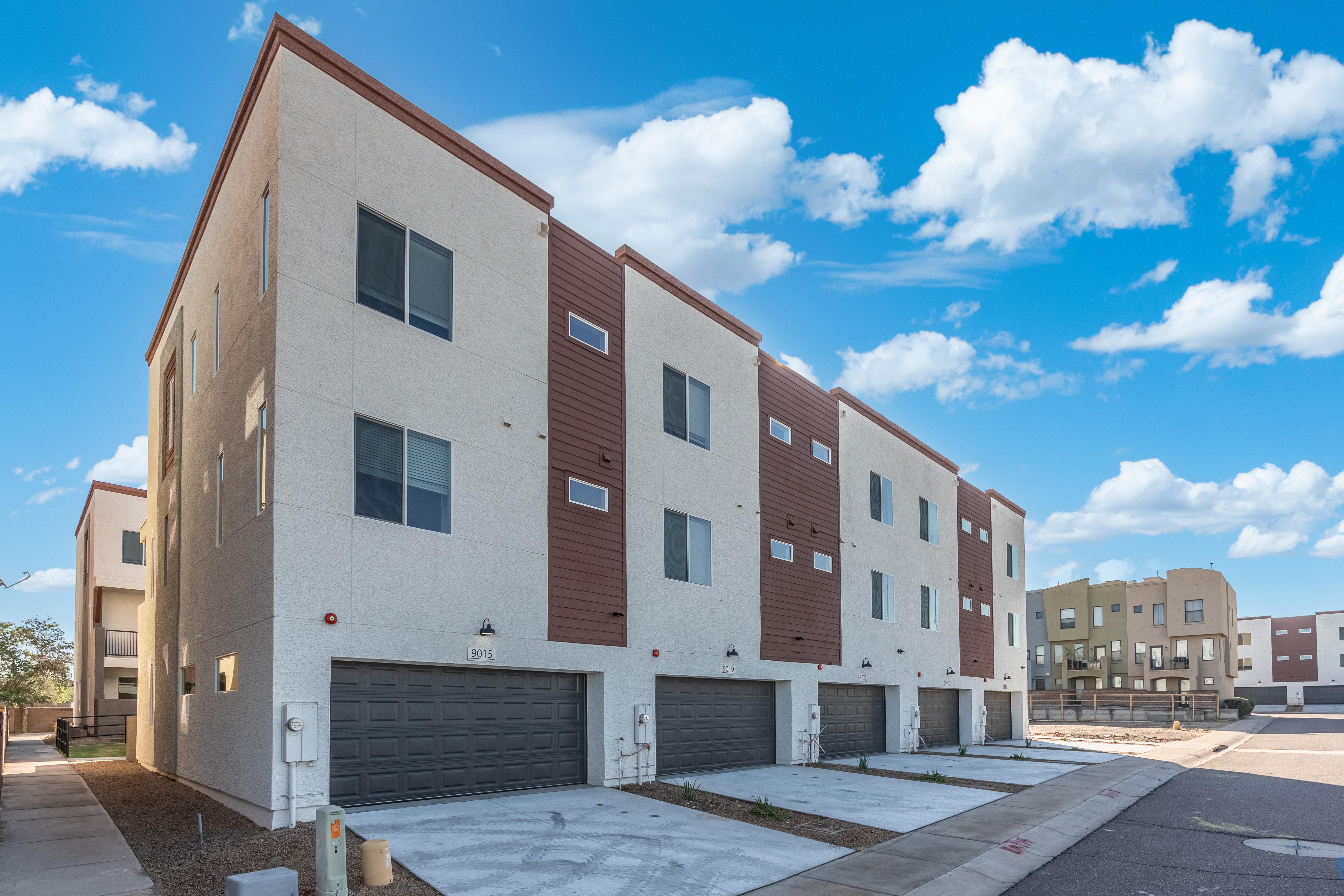 A row of contemporary townhouses with garages, featuring a mix of white and brown exterior finishes under a bright blue sky with fluffy clouds.