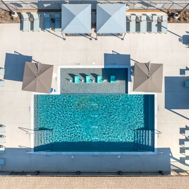 Aerial view of a swimming pool area featuring a rectangular pool surrounded by lounge chairs and umbrellas, with landscaped edges and a clean, modern design.