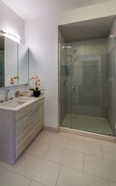 A modern bathroom featuring a glass shower enclosure, light-colored tiles, a wooden vanity with a sink, and a wall-mounted mirror. Flowers are placed on the countertop.