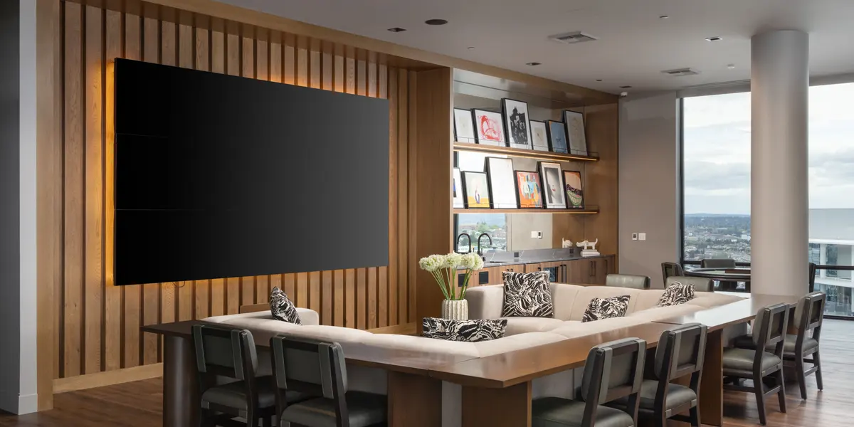 A modern living room with large windows offering a view, featuring a long rectangular table surrounded by dining chairs. A large flat-screen TV is mounted on a wooden wall, and shelves with framed pictures are visible in the background. Decorative pillows are placed on a sectional couch.