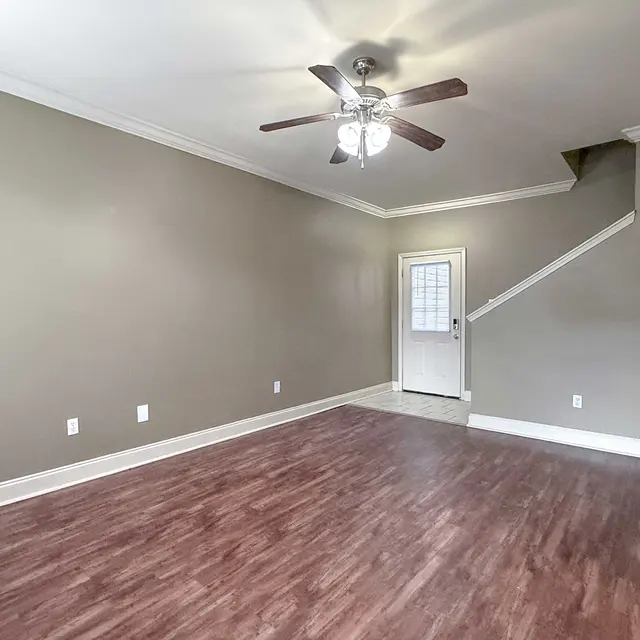 A spacious empty living room with light brown walls and a dark hardwood floor. A ceiling fan is visible, and a staircase leads to an upper level on the right side of the image. A door is located in the center background, providing access to the outside.