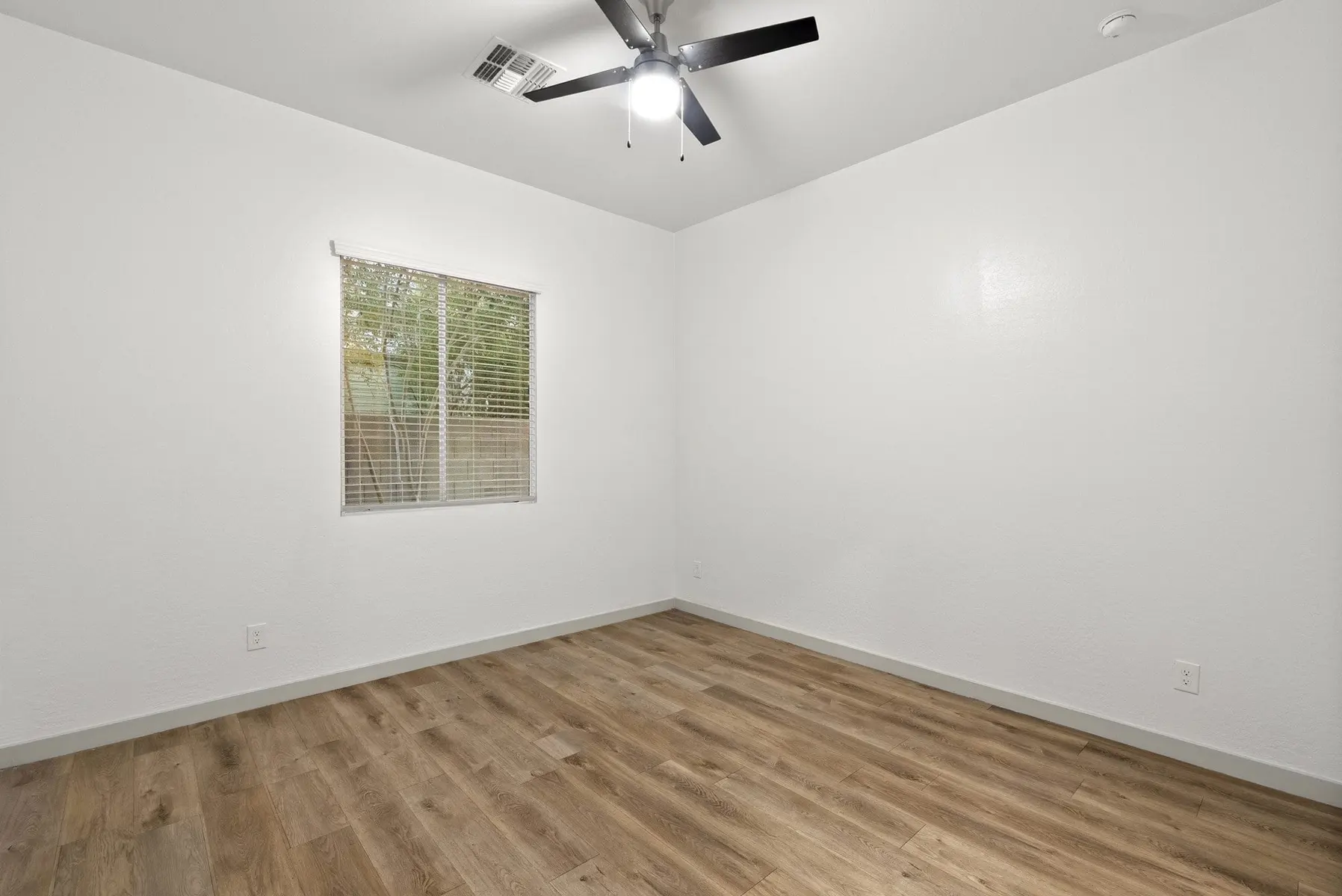 A minimalist empty room with a wooden floor, white walls, a ceiling fan, and a window with blinds.
