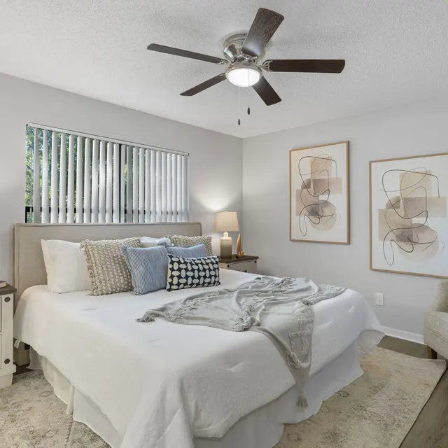A modern bedroom featuring a bed with decorative pillows and a throw blanket, a nightstand with a lamp, a fan overhead, and two abstract art pieces on the wall.