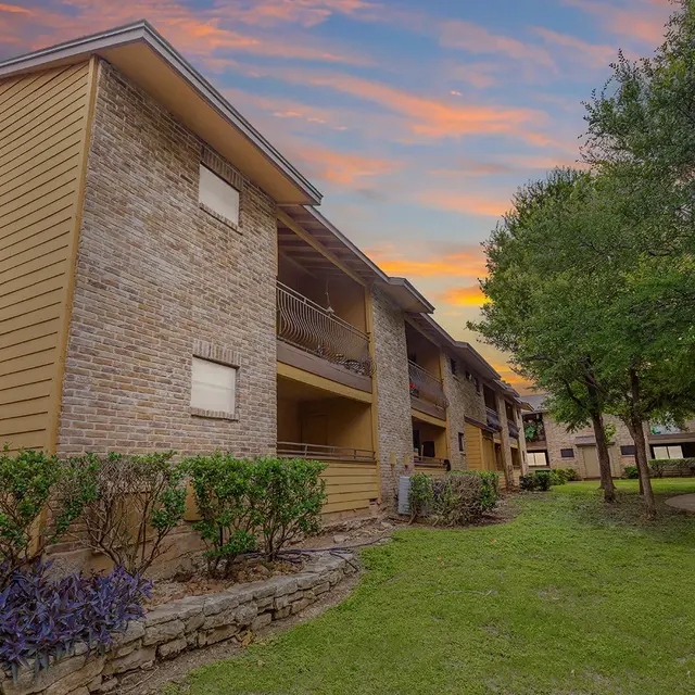 An apartment complex featuring two-story buildings with a mix of brick and wooden siding, surrounded by well-maintained greenery and shrubs, under a colorful sunset sky.