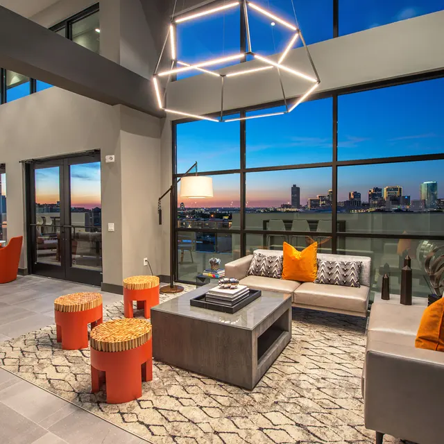 Stylish Modern Living Room with City View A modern living room with large windows showcasing a city view at dusk. The space features contemporary furniture, including a gray sofa, orange accent cushions, a coffee table, and round stools. A geometric light fixture hangs from the ceiling, adding a stylish touch.