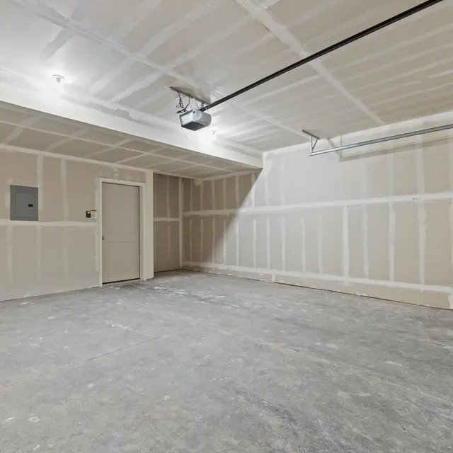An empty garage with unfinished walls and concrete floor, featuring a garage door, side entry door, and minimal lighting.