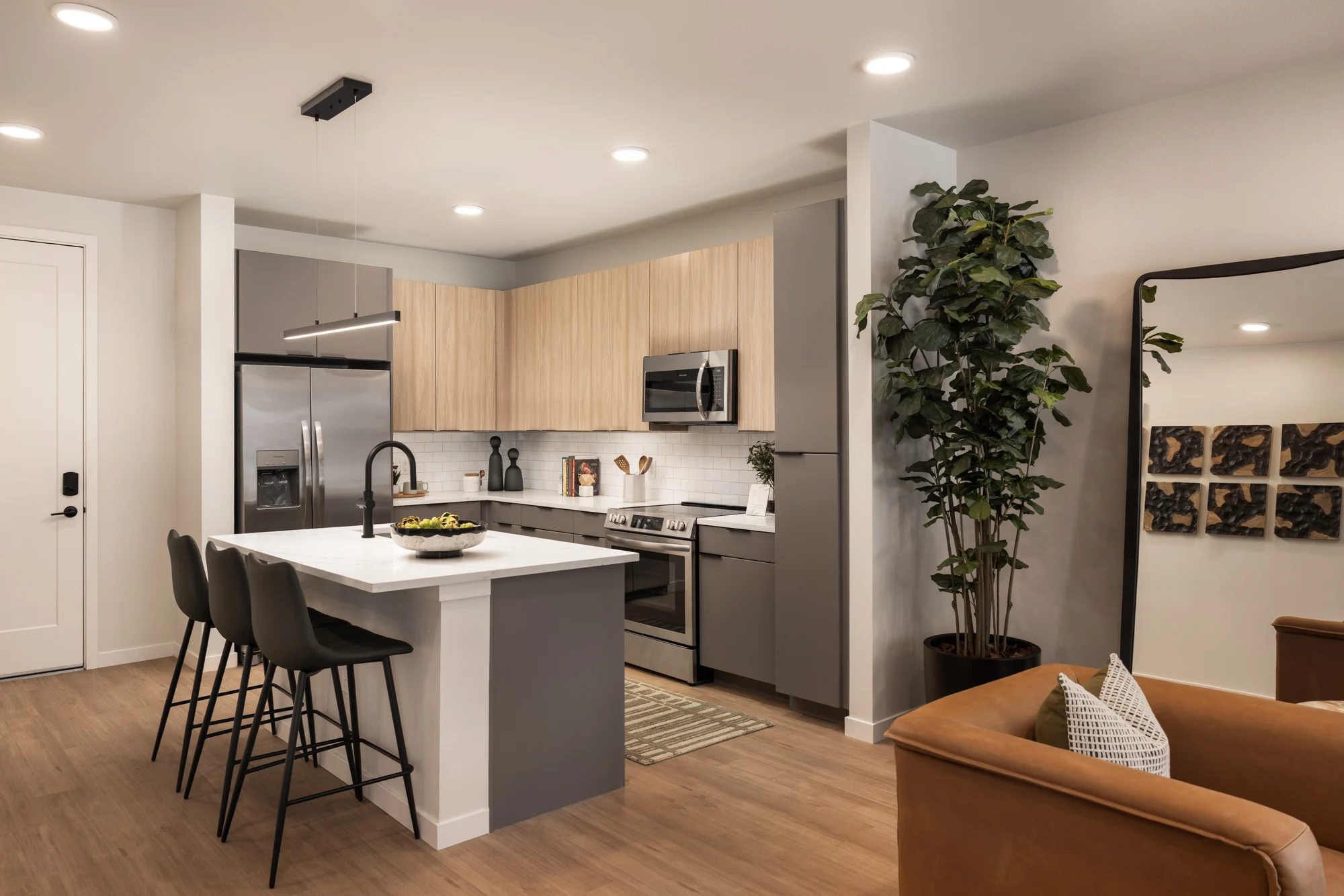A modern kitchen with a central island, bar stools, cabinetry, and stainless steel appliances. There are plants, a mirror, and decorative art in the living space.