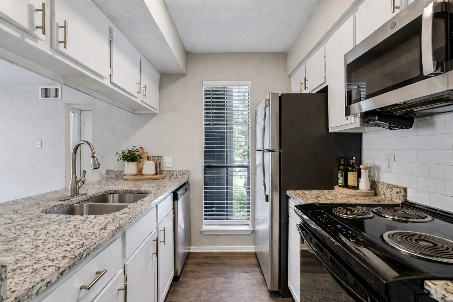 A modern kitchen featuring a granite countertop, white cabinetry, stainless steel appliances, and a large window with blinds.