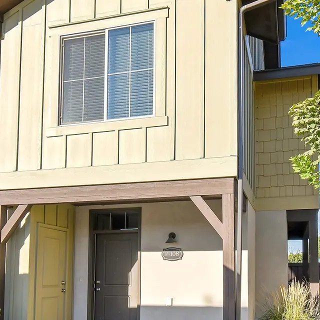 A two-story house with a light green facade and a brown entryway. It features a large window on the upper floor and a porch area below. The house is surrounded by greenery and is part of a residential neighborhood.