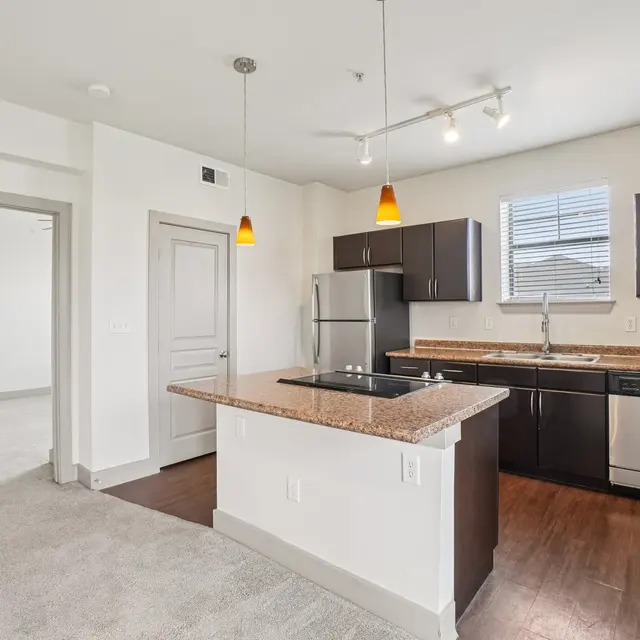 A modern kitchen featuring dark cabinetry, a granite countertop island, stainless steel appliances, and pendant lighting. The room is open and bright, with a window allowing natural light.
