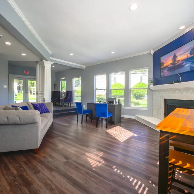 A stylish modern living area featuring a gray couch with purple throw pillows, a wooden table with chairs, and a bright atmosphere with large windows.