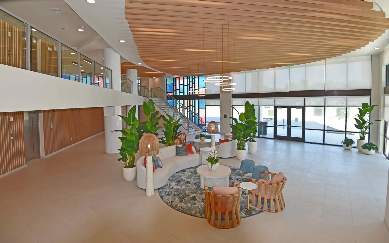 A spacious and modern lobby featuring a comfortable seating area with a circular couch, colorful chairs, potted plants, and decorative light fixtures. Large windows provide ample natural light, and a staircase is visible in the background.